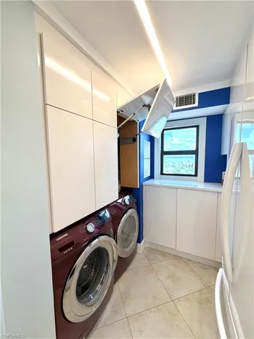 Modern laundry room featuring front-load washer/ dryer in rich burgundy finish, tile flooring, recessed ceiling lighting, white cabinetry with upper storage, and a blue accent wall with windows for natural light