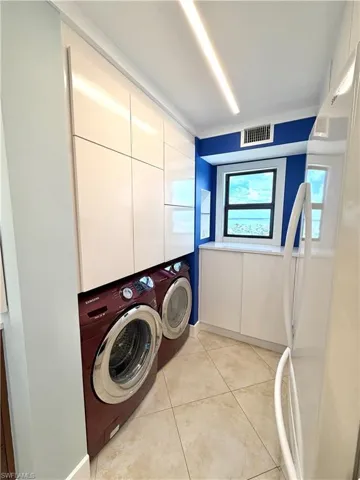 Modern laundry room featuring side-by-side front-load washer/dryer with burgundy cabinetry, tile flooring, recessed ceiling lighting, and abundant white cabinetry storage