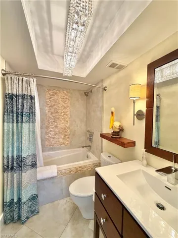 Recessed ceiling lighting with a crystal chandelier, tiled bathtub surround with an integrated shower, wall-mounted floating shelf, vanity with an integrated sink, and a wood-framed mirror