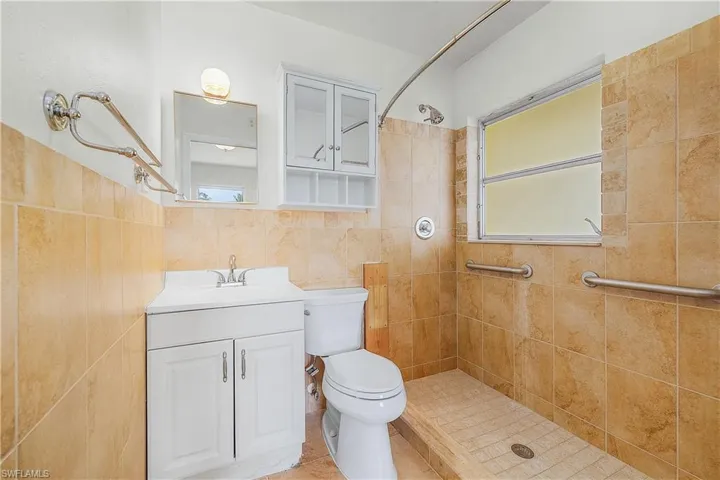 Bathroom with tiled shower, vanity, toilet, tile walls, and plenty of natural light