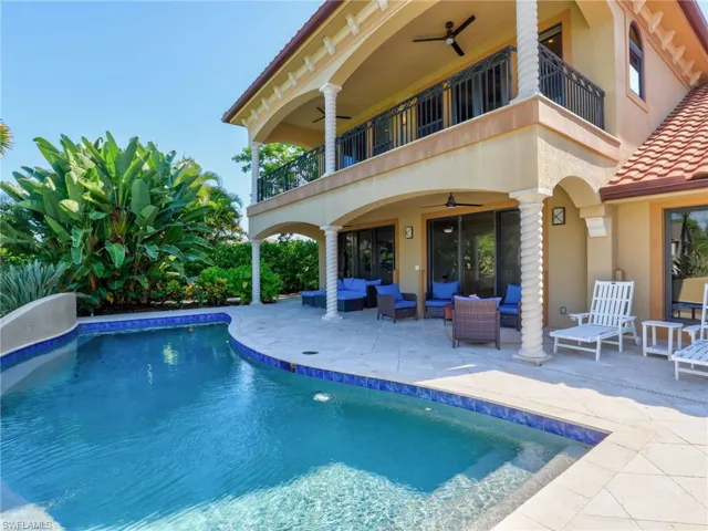 Luxurious resort-style backyard oasis featuring a sparkling saltwater pool with integrated spa, travertine decking, and a two-story Mediterranean-inspired residence with covered loggia, ceiling fans, and French doors opening to the outdoor entertaining space