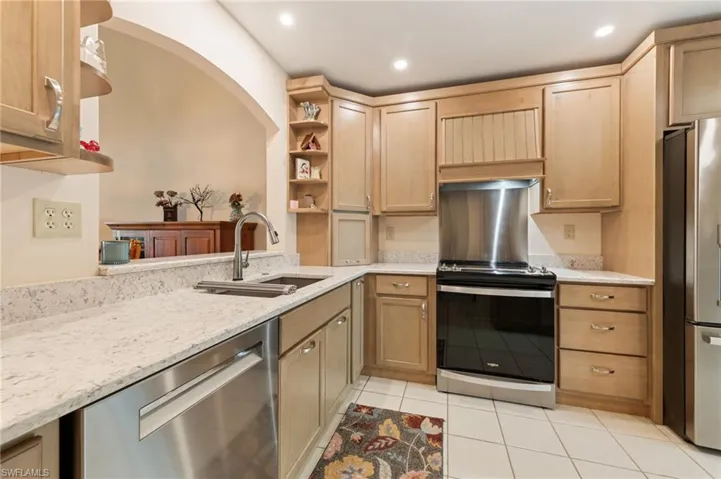 Kitchen featuring natural wood cabinetry, light-toned stone-finish countertops, stainless steel appliances, a stainless steel undermount sink with a gooseneck faucet, and tiled flooring