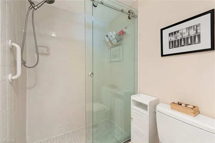 Walk-in shower enclosure with a sliding glass door, white tiled walls, adjustable handheld shower fixture, and wall-mounted support bar