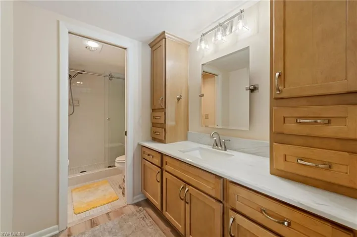 Bathroom featuring wood-finish cabinetry, stone-surface countertop with an integrated sink, glass-enclosed shower with a handheld fixture, and wall-mounted vanity lighting