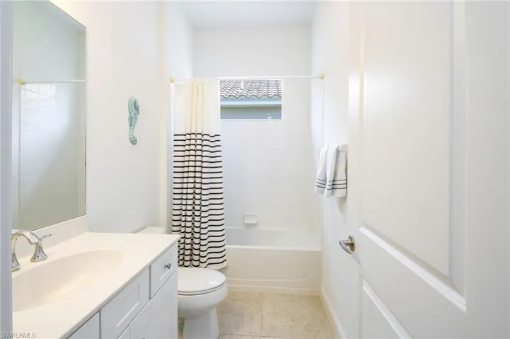 Carpeted bedroom featuring a tray ceiling with a ceiling fan, three windows with white plantation shutters, an open doorway revealing a bathroom with a vanity and shower, and a second open doorway