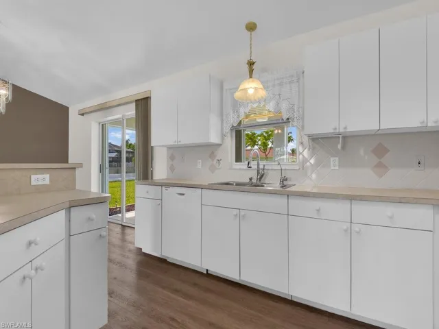 Kitchen featuring white cabinetry, light-toned countertops, a white tile backsplash with accent squares, stainless steel sink, and wood-finish flooring