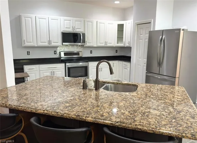 Kitchen featuring an island with a built-in stainless steel sink, light-toned stone countertops, white cabinetry, dark-toned countertops, and stainless steel appliances