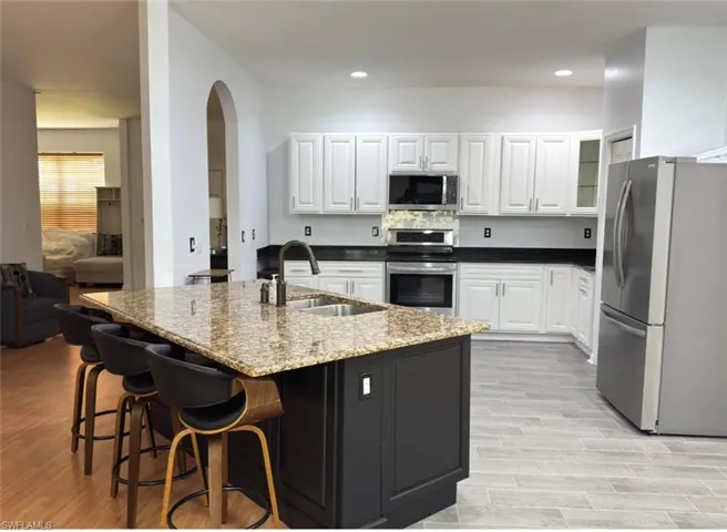 Kitchen featuring an arched doorway, white cabinetry, dark countertops, a central island with a sink, and stainless steel appliances
