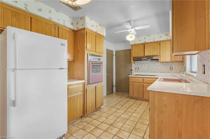Kitchen featuring ceiling fan, light tile patterned floors, sink, white appliances, and exhaust hood, built in over and solid wood cabinets, cook top and double sinks with window looking out to front yard, door to garage, pantry and door to the dining area of great room
