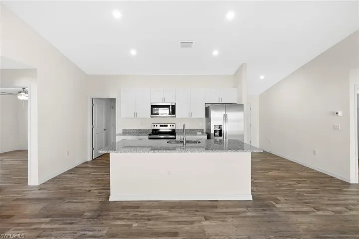 Kitchen with white cabinets, appliances with stainless steel finishes, light stone counters, a center island with sink, and dark wood-style floors