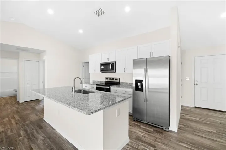 Kitchen featuring appliances with stainless steel finishes, vaulted ceiling, light stone counters, white cabinets, and an island with sink
