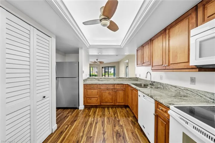 Kitchen with brown cabinetry, a ceiling fan, dark wood-style floors, a tray ceiling, and white appliances