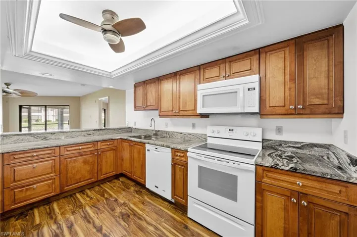 Kitchen with white appliances, brown cabinetry, a raised ceiling, dark wood-style floors, and crown molding