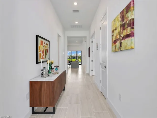 Recessed lighting illuminates a hallway with light-toned tile flooring and white walls, leading to an open living area with sliding glass doors
