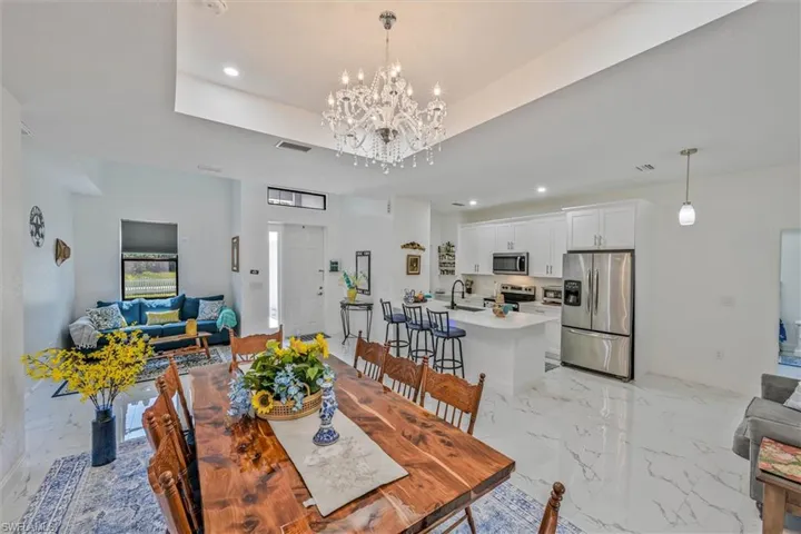 Open-concept living space featuring a tray ceiling with a crystal chandelier, wood-finish dining table, light-toned flooring, and a contemporary kitchen with white cabinetry and stainless steel appliances