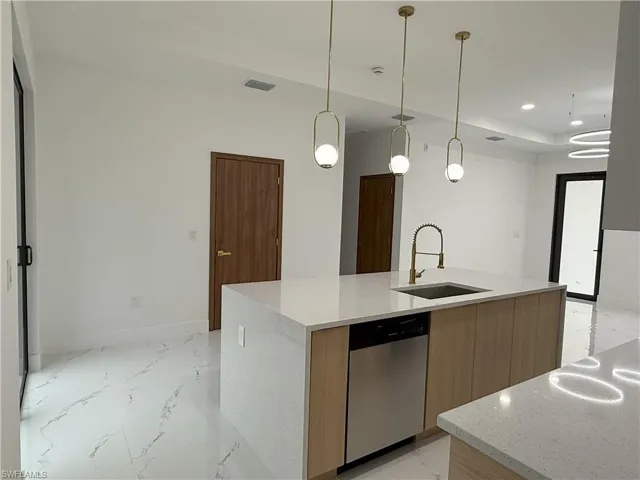 Kitchen with pendant lighting, light stone counters, dishwasher, light marble finish floors, and an island with sink