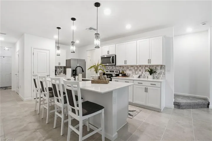 Modern kitchen featuring a white center island with integrated seating, stainless steel appliances, white cabinetry, and an upgraded geometric tile backsplash.  Upgraded pendant lighting fixtures.