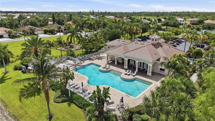 Community clubhouse with a barrel-tile roof, featuring a large outdoor swimming pool with integrated steps, surrounding paved deck, and a lush, tropical landscape