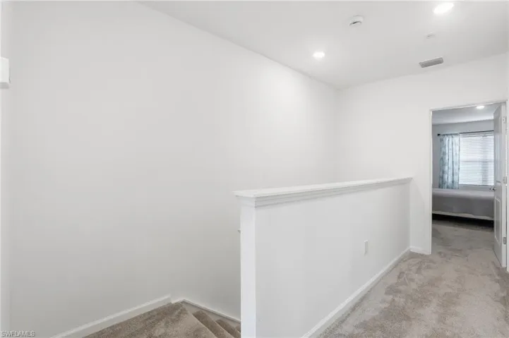 Upper-level hallway featuring plush gray carpeting, recessed ceiling lighting, a white wall-mounted handrail, and a doorway leading to an additional space