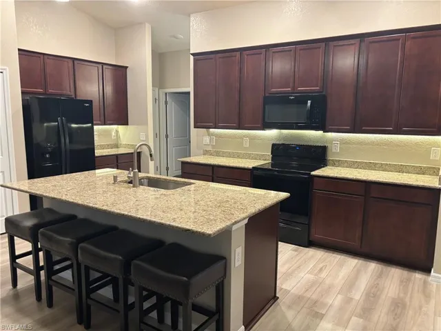 Kitchen with black appliances, light stone counters, light wood finished floors, a kitchen bar, and a kitchen island with sink