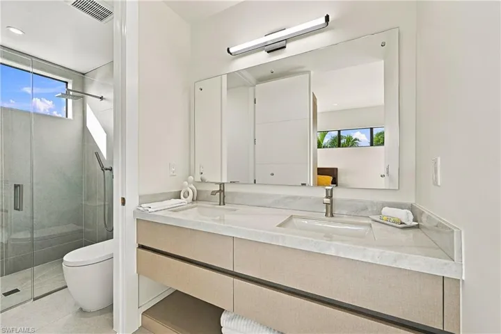 Double vanity featuring a stone countertop, under-mount sinks, wall-mounted brushed-metal faucets, a frameless wall-to-wall mirror, and a walk-in glass-enclosed shower with tiled walls