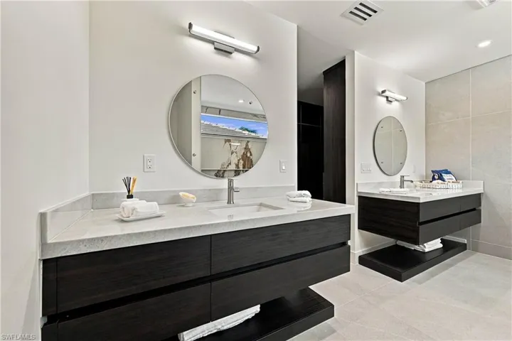 Dual floating wood-finish vanities featuring light-toned stone countertops, rectangular undermount sinks, circular wall-mounted mirrors, and sleek horizontal light fixtures