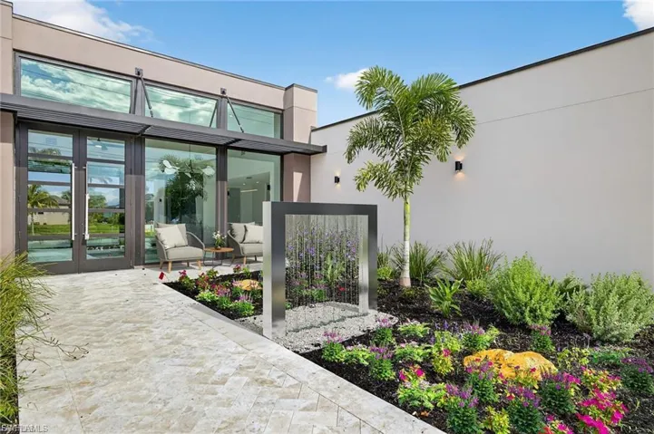 Modern exterior entrance featuring floor-to-ceiling glass paneling, a structured metal water feature, herringbone-patterned stone pavers, and a landscaped garden bed