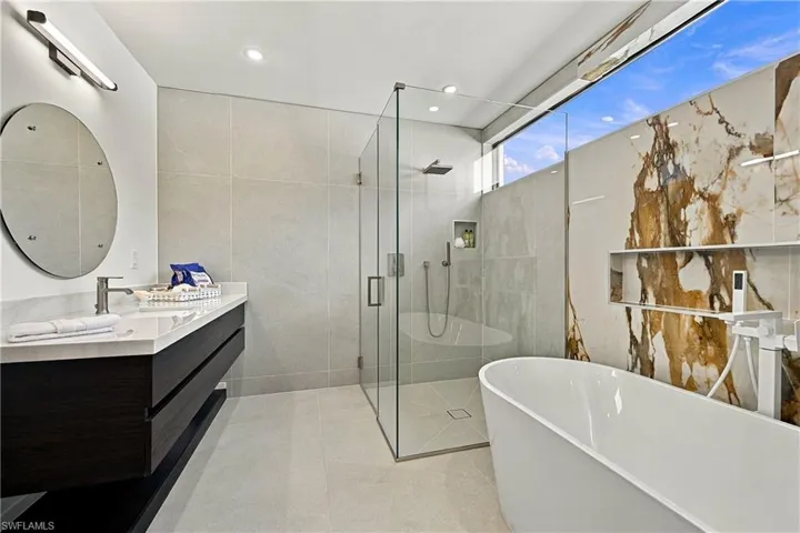 Floating vanity with rectangular countertop, glass-enclosed shower, stone wall accent, freestanding soaking tub, and clerestory window
