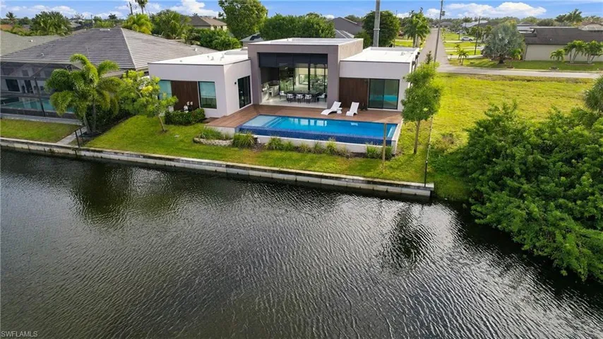 Modern waterfront residence featuring a flat-roof architectural design, a rectangular pool with a tile surround, an expansive wood-finish deck, and extensive glass-paneled exterior walls
