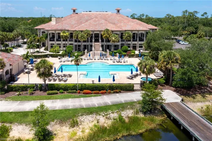 Expansive clubhouse featuring a multi-level architectural design, clay tile roof, large swimming pool with surrounding paver deck, and a wooden pedestrian bridge