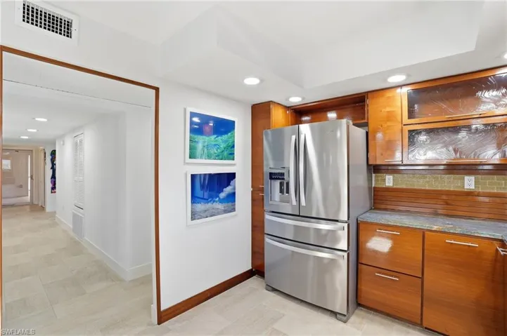 Kitchen featuring brown cabinetry, dark stone countertops, stainless steel fridge with ice dispenser, recessed lighting, and glass insert cabinets - Virtually Edited Image