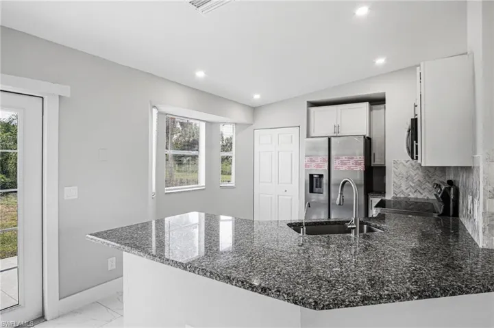 Kitchen featuring a large island with a stainless steel sink and a high-arc faucet, granite countertops, an integrated stainless steel refrigerator, white cabinetry, and a herringbone-patterned tile backsplash