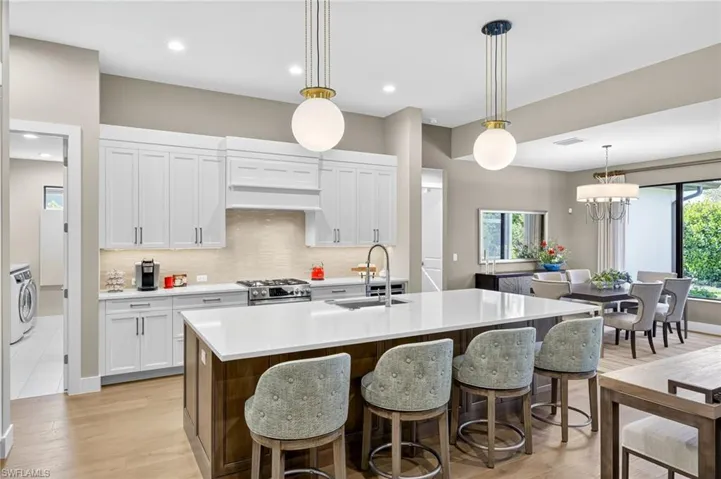 Kitchen with tasteful backsplash, light countertops, light wood floors, expanded kitchen island with sink, and white custom cabinets and additional custom storage