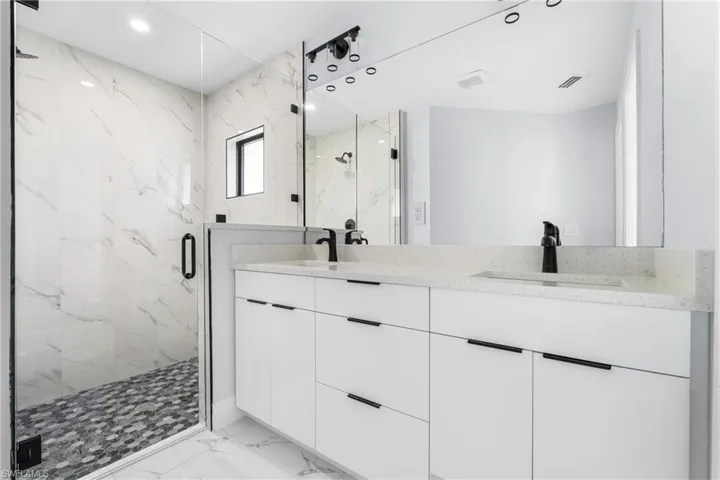 Spacious bathroom featuring a frameless glass shower enclosure with stone mosaic flooring, dual white vanities with integrated sinks, and black hardware throughout