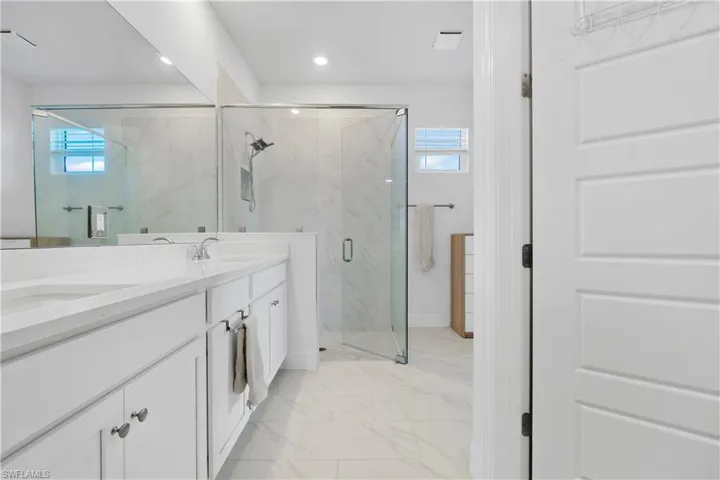 Dual-sink vanity with white cabinetry and countertop, glass-enclosed walk-in shower with tiled surround, large wall-to-wall mirror, and oversized floor tiles