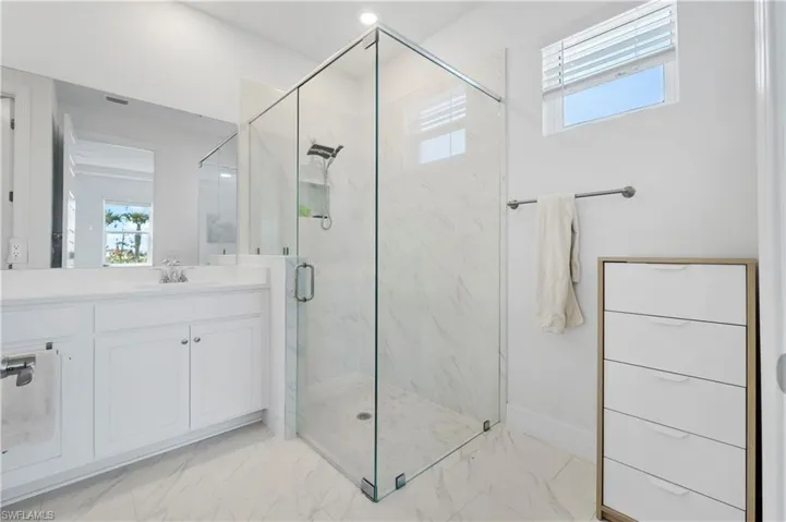 Walk-in shower with a glass enclosure, wall-mounted showerhead, light-toned stone-finish tiling, white cabinetry vanity with a countertop basin, and large wall-mounted mirror