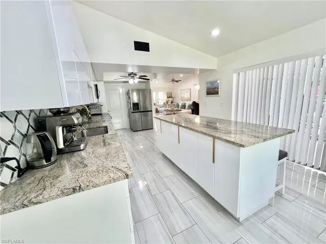 Modern kitchen featuring a spacious island with a light-toned granite countertop, white cabinetry with linear gold-toned pulls, a geometric tile backsplash, and large format tile flooring