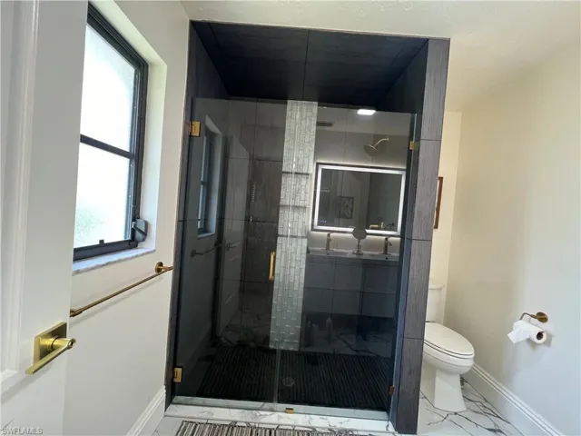 Modern bathroom featuring a frameless glass shower enclosure with brass hardware, a dark-finish vanity with dual sinks, an illuminated mirror, and marble-look floor tiling
