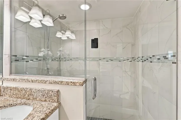 Shower stall featuring full-height tile surround with a glass door, decorative mosaic tile inlay, recessed ceiling lighting, and a stone-topped vanity