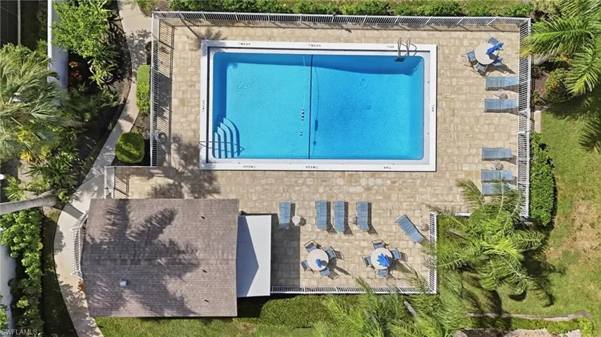 Rectangular in-ground swimming pool with integrated stair entry, surrounded by paved decking, enclosed by white metal perimeter fencing, and featuring a covered patio structure