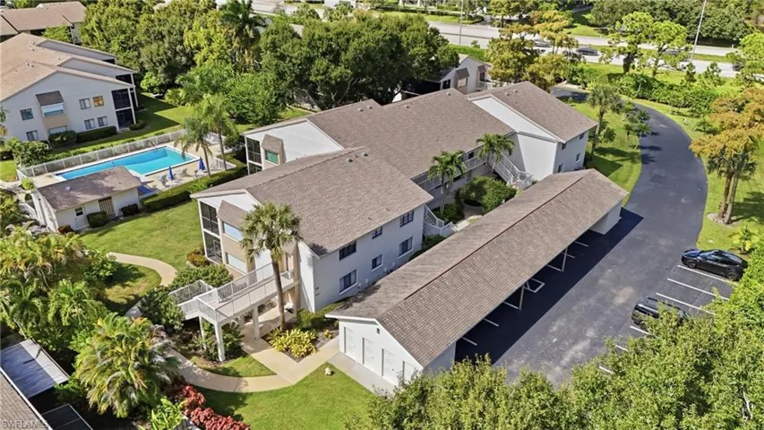 Multi-story building complex featuring a shingled roof, ground-level covered parking, manicured landscaping with mature palm trees, and an outdoor swimming pool