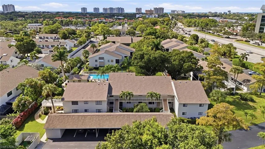 Aerial perspective of multi-family complex featuring a shingled roof, attached covered parking, communal swimming pool, mature palm trees, and manicured grounds