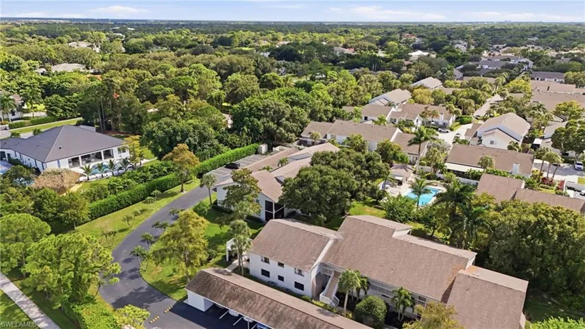 Aerial perspective of multi-family residential structures featuring shingle roofing, paved surface parking, and dense mature tree coverage