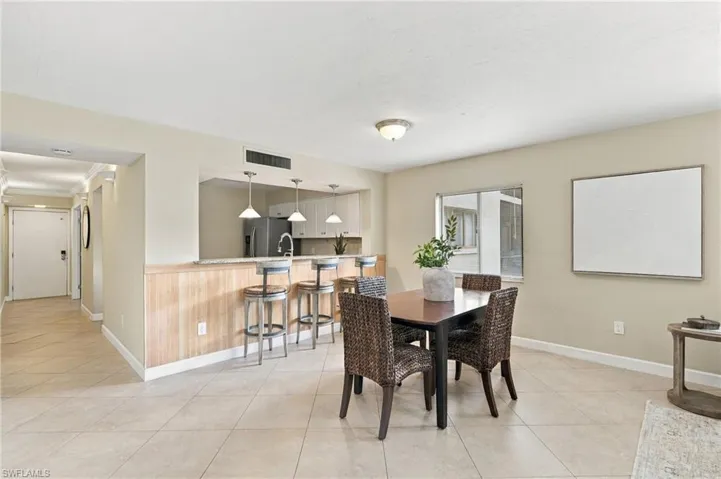 Open-concept floor plan featuring light-toned tile flooring, pass-through kitchen window with breakfast bar seating, wood-paneled bar front, and three pendant light fixtures