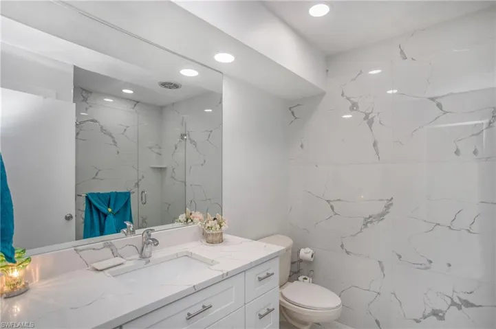 Recessed lighting illuminating a vanity with a white countertop and an integrated sink, complemented by a full-width mirror and floor-to-ceiling tile in a marbled pattern