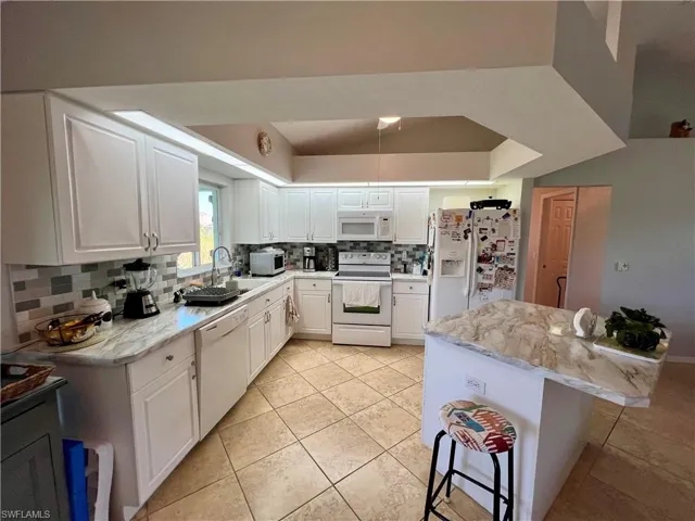 Kitchen featuring a kitchen bar, decorative tiled backsplash, light stone counters, white appliances, and white cabinetry