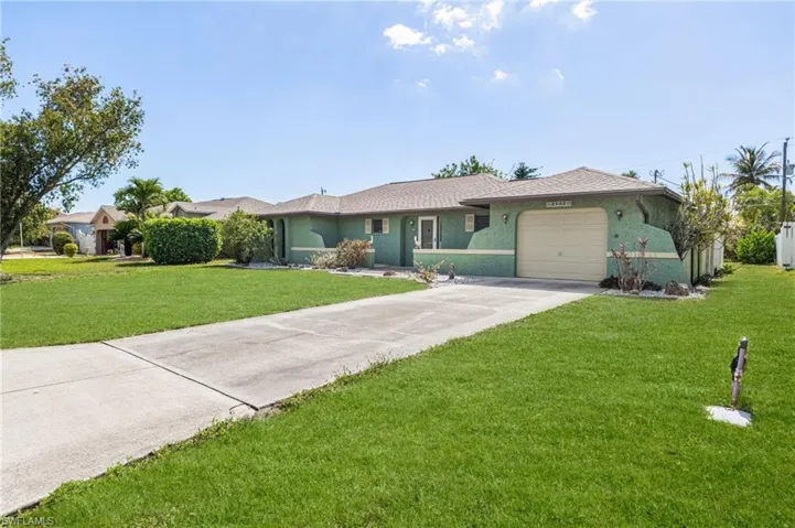 Single-story residence featuring a turquoise stucco exterior, a tan garage door, a concrete driveway, a front lawn, and mature landscaping