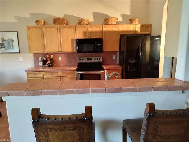 Kitchen with backsplash, tile counters, black appliances, light brown cabinetry, and kitchen peninsula