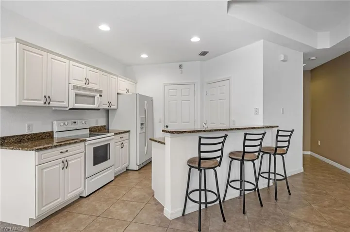 Kitchen featuring white cabinets, kitchen peninsula, dark stone counters, white appliances, and light tile patterned floors