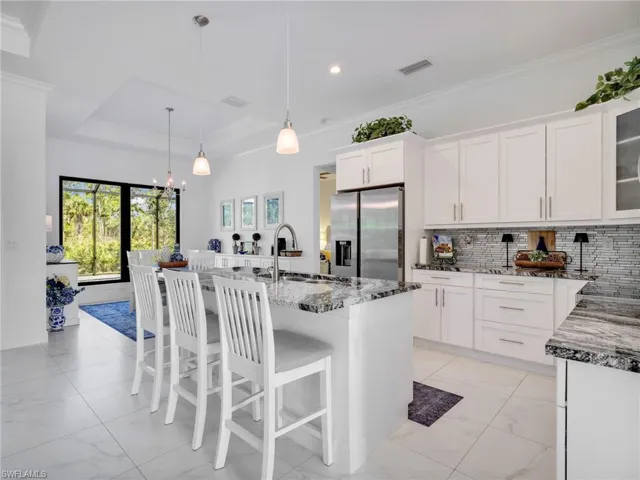 Kitchen With White Shaker Cabinetry, Island, Pendant Lighting, Glass Tile Backsplash, Stainless-Steel Appliances, Quartz Counters, & Dining Area With Built-In Bar and Quartz Counter For Extra Storage With Pool & Wooded Rear Yard Views
